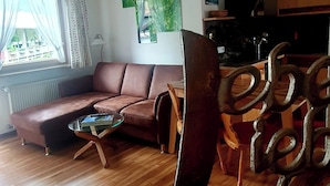 Un salón con un sofá de cuero marrón, una mesa de centro de vidrio y una mesa de comedor de madera con sillas de metal. Hay una ventana con cortinas, un radiador y un cuadro abstracto verde en la pared.