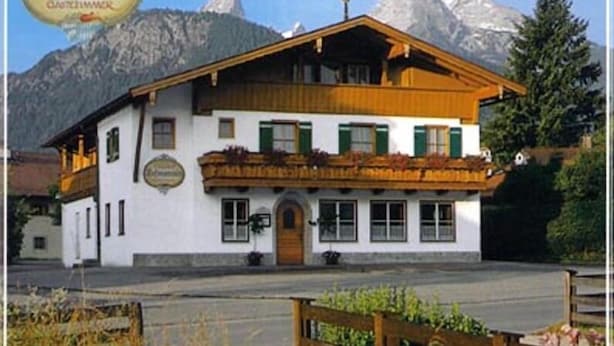 Un edificio de estilo alpino tradicional con un balcón de madera, un cartel que indica que es un Gasthof, y montañas al fondo.