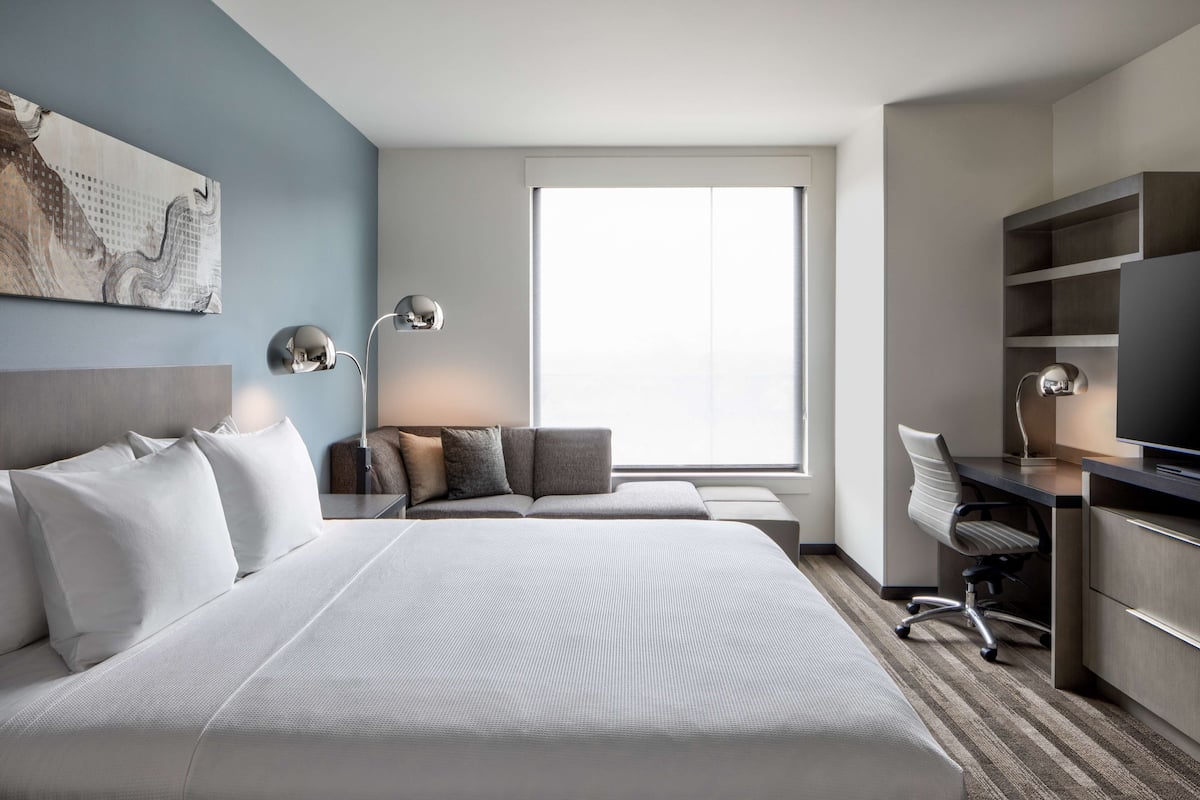 A hotel room with a large bed, a desk with a chair, a television, and a large window.