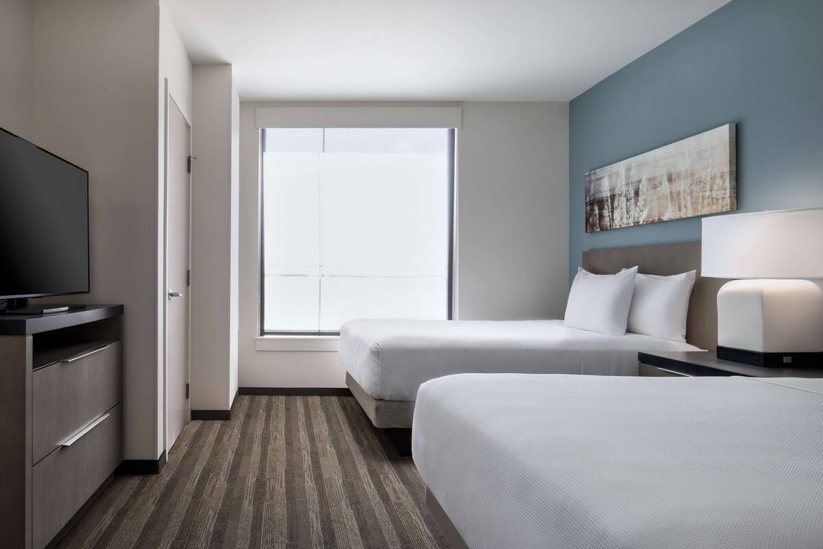 A hotel room with two beds, a television, a desk with a lamp, and a large window.