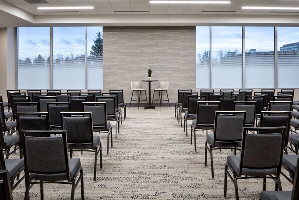 A conference room with rows of chairs facing a stage, large windows, and a carpeted floor.