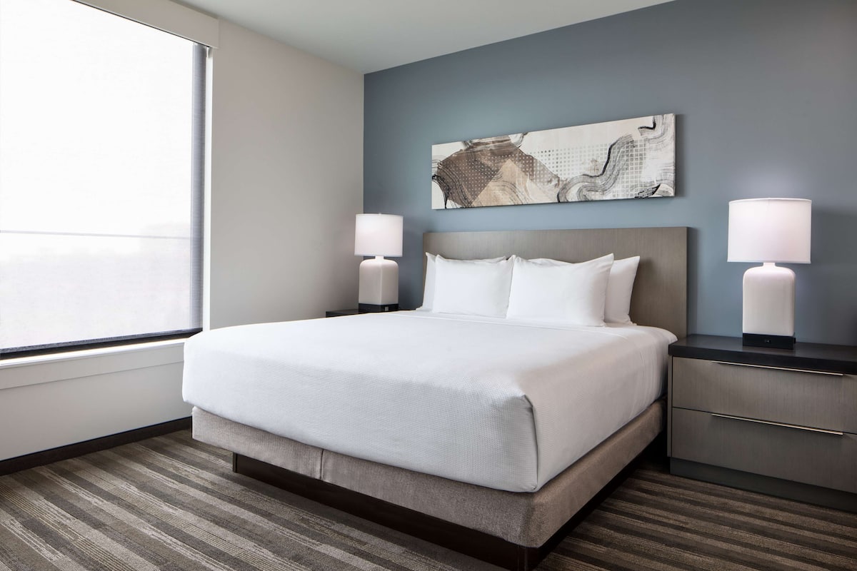 A hotel room with a large bed, two bedside tables with lamps, a wall-mounted artwork, and a window providing a view.