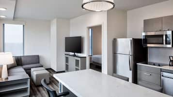 A modern hotel room with a kitchenette, a flat-screen TV, a sofa, and a dining area.