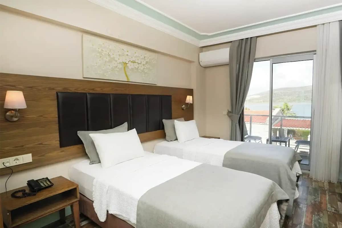 A hotel room with two beds, a wooden headboard, a painting, an air conditioning unit, a balcony with a view, and a desk with a kettle.