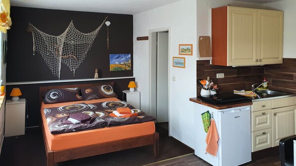Une pièce de taille modeste comprenant un lit, un petit coin cuisine et un mur décoré d’une œuvre en macramé et d’autres œuvres d’art.