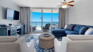 A living room with a blue sofa, a television, a ceiling fan, and a view of the ocean through large sliding doors.