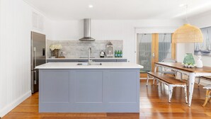 A modern kitchen with a central island, a stainless steel range hood, and a dining area with a wooden table and chairs. - Sea Deck, Hyams Beach, Jervis Bay (Hyams Beach)