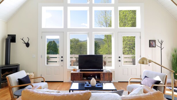 Een ruime woonkamer met een grote televisie, zachte zithoek, een open haard en grote ramen met uitzicht op groen.