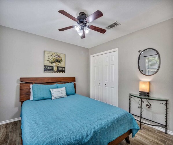 A bedroom with a bed, a ceiling fan, a door, a mirror, and a small table with a lamp.