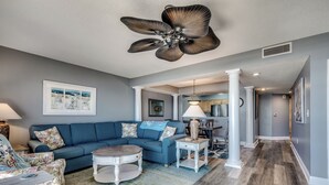 A living room with a blue sofa, a round coffee table, and a ceiling fan. There is a kitchen area in the background with a dining table and chairs. - 🌞🌊Lazy River + Indoor/Outdoor Pools | Oceanfront | South Wind 402 (Myrtle Beach)