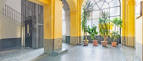 Um espaço amplo e interno com piso ladrilhado, paredes amarelas e janelas grandes. Há plantas em vasos e uma escada com corrimão de metal.