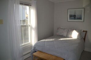 A bedroom with a bed, a window with sheer curtains, a framed picture on the wall, and a wooden bench.