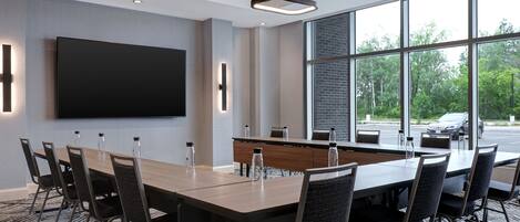 A conference room with a long table, chairs, a flat-screen TV, and large windows offering a view of the outdoors.