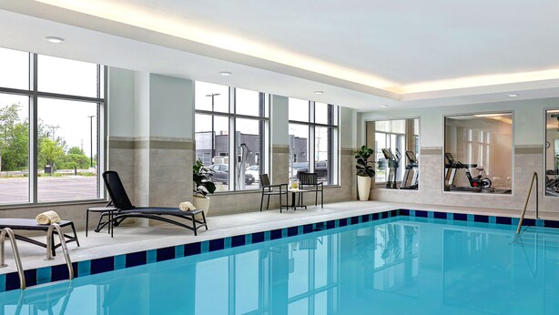 Piscina de hotel con sillas de descanso, equipo de ejercicio y ventanas amplias que dan vista al estacionamiento.