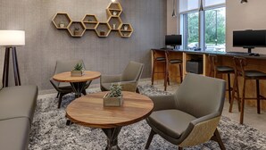 Un espacio de oficina moderno con una mesa redonda de madera, dos sillas, un estante hexagonal con plantas, un escritorio con una computadora y un ventanal con vistas a árboles y coches.