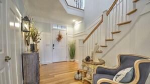 A spacious hallway with a wooden floor, a staircase, a grey armchair, a small table with decorative items, and a door with a wreath. - "Haven By The Sound" - Intercoastal Waterway Home w/ Pool, 6 Bedrooms / 6.5 Bathrooms (Holden Beach)