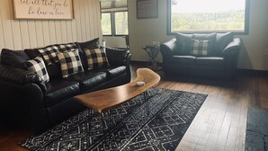 A living room with two black leather sofas, a wooden coffee table, and a patterned rug. A framed quote is on the wall, and there is a window with a view of greenery. - Camp HighBridge 2 Bed 1 Bath Big Manistee River, Private Frontage. Bunk House! (Wellston)