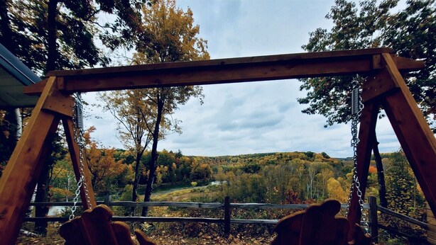 A wooden swing set with two chairs and a table, overlooking a forested valley with a river. - Camp HighBridge 2 Bed 1 Bath Big Manistee River, Private Frontage. Bunk House! (Wellston)