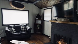 A cozy living room with a fireplace, a black leather sofa, and a television above the fireplace. - Camp HighBridge 2 Bed 1 Bath Big Manistee River, Private Frontage. Bunk House! (Wellston)