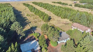 Vue aérienne d’une propriété située sur la côte, comprenant une grande maison, un bâtiment plus petit et une zone boisée avec un sentier de terre.
