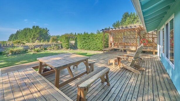 Une terrasse en bois avec des tables de pique-nique et des bancs, une pergola agrémentée de décorations suspendues, et une vue sur une zone boisée.
