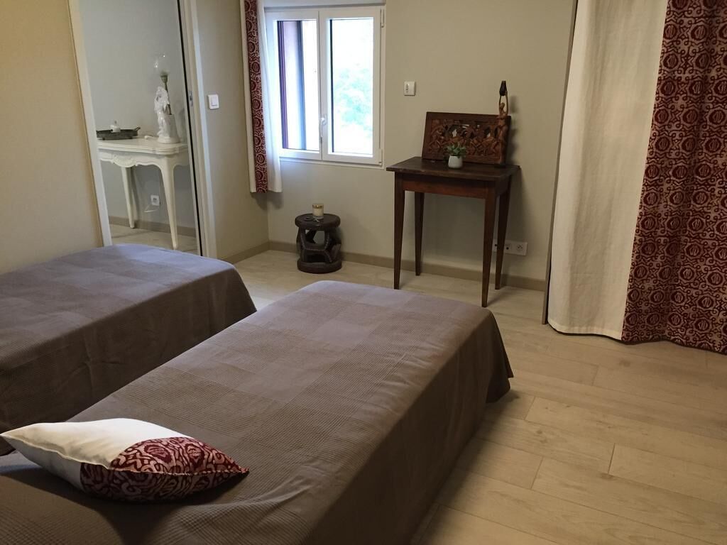 Een kamer met twee bedden, een klein tafeltje met een fotolijstje, een beeld en een raam met gordijnen.
