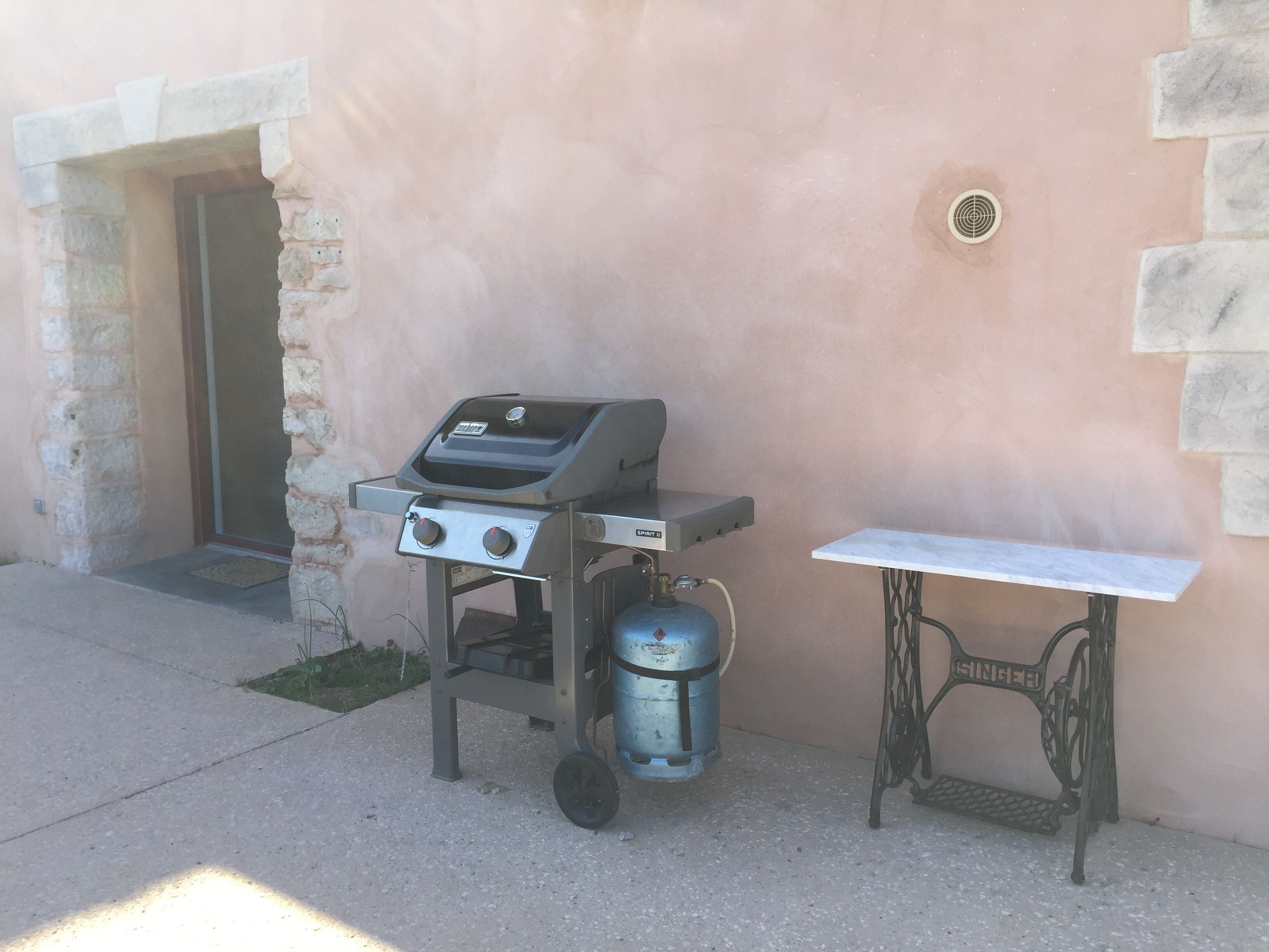 Una parrilla portátil con un tanque de gas y una mesa metálica al lado de un muro de piedra.