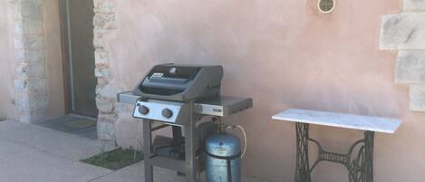 Una parrilla portátil con un tanque de gas y una mesa metálica al lado de un muro de piedra.
