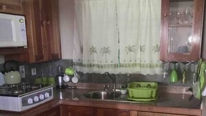 A compact kitchen with wooden cabinets, a gas stove, a microwave, and a window with a curtain. - 1 Bed Villa - Sleeps 2 - Garden - Parking - Wifi (Ocho Rios)