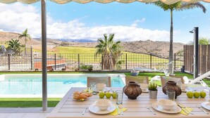 Un espacio de comedor al aire libre con vista a una piscina, una mesa dispuesta para dos personas y un paisaje pintoresco al fondo.