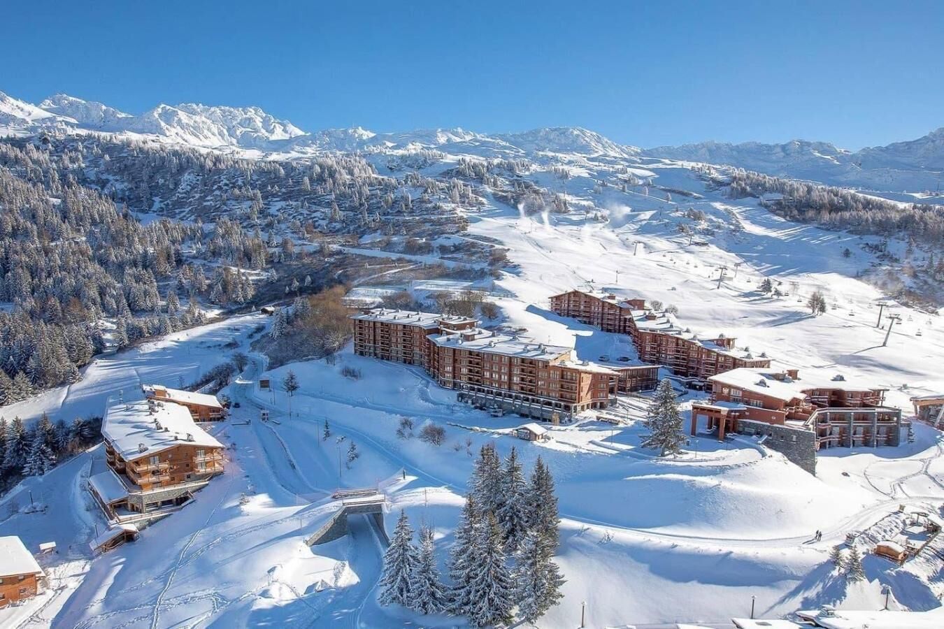 Une station de montagne enneigée, avec plusieurs bâtiments, des pistes de ski et un ciel dégagé.