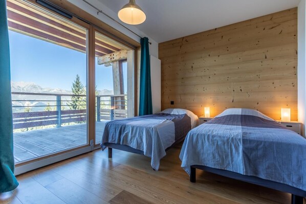 Two single beds with matching headboards, a balcony with mountain views, and a warm, natural ambiance.