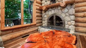 Interior d'una cabana de fusta amb una llar de pedra, una taula de fusta gran i una finestra que dona a un bosc.