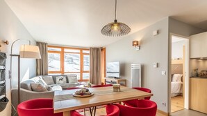 A modern living room with a dining area, large windows, a flat-screen TV, and a kitchen visible through an open door. - Ski-in/ski-out & foot of the slopes, 7th floor, view ski slopes, fitness, swimming pool, sauna (Bourg-Saint-Maurice)