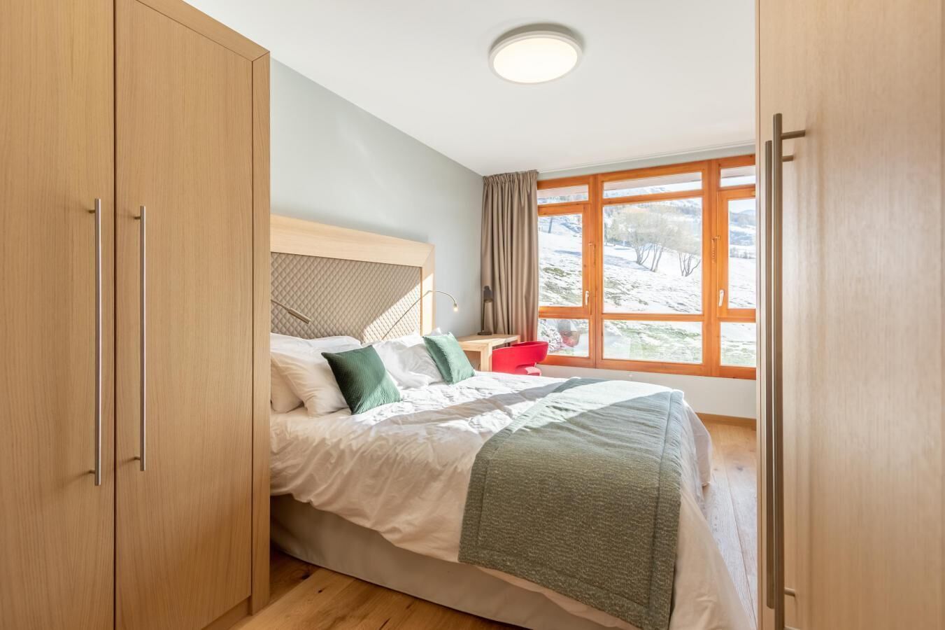 A bedroom with a bed, wooden wardrobe, and a window with a view of a snowy landscape.