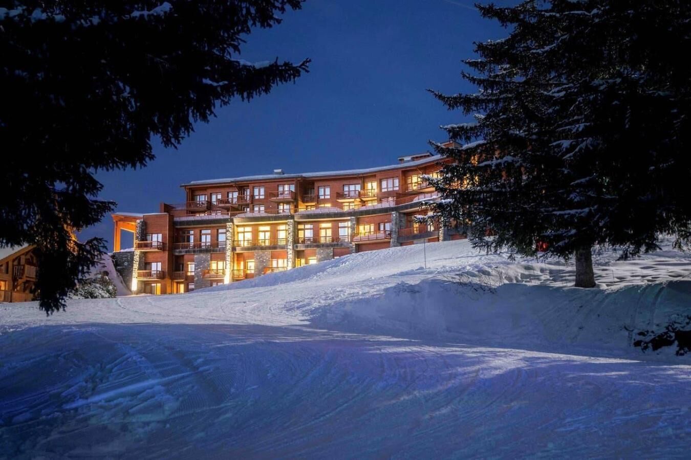 A multi-story hotel with a ski slope in the foreground, illuminated windows, and snow-covered surroundings.