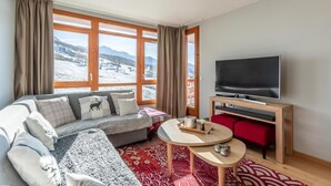 A cozy living room with a sofa, a flat-screen TV, and a view of a snowy landscape through large windows. - Ski-in/ski-out & foot of the slopes, 7th floor, view ski slopes, fitness, swimming pool, sauna (Bourg-Saint-Maurice)