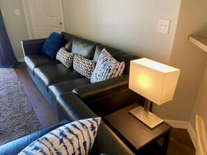 A living room with a black leather sofa, patterned cushions, a small wooden side table with a lamp, and a carpeted floor.