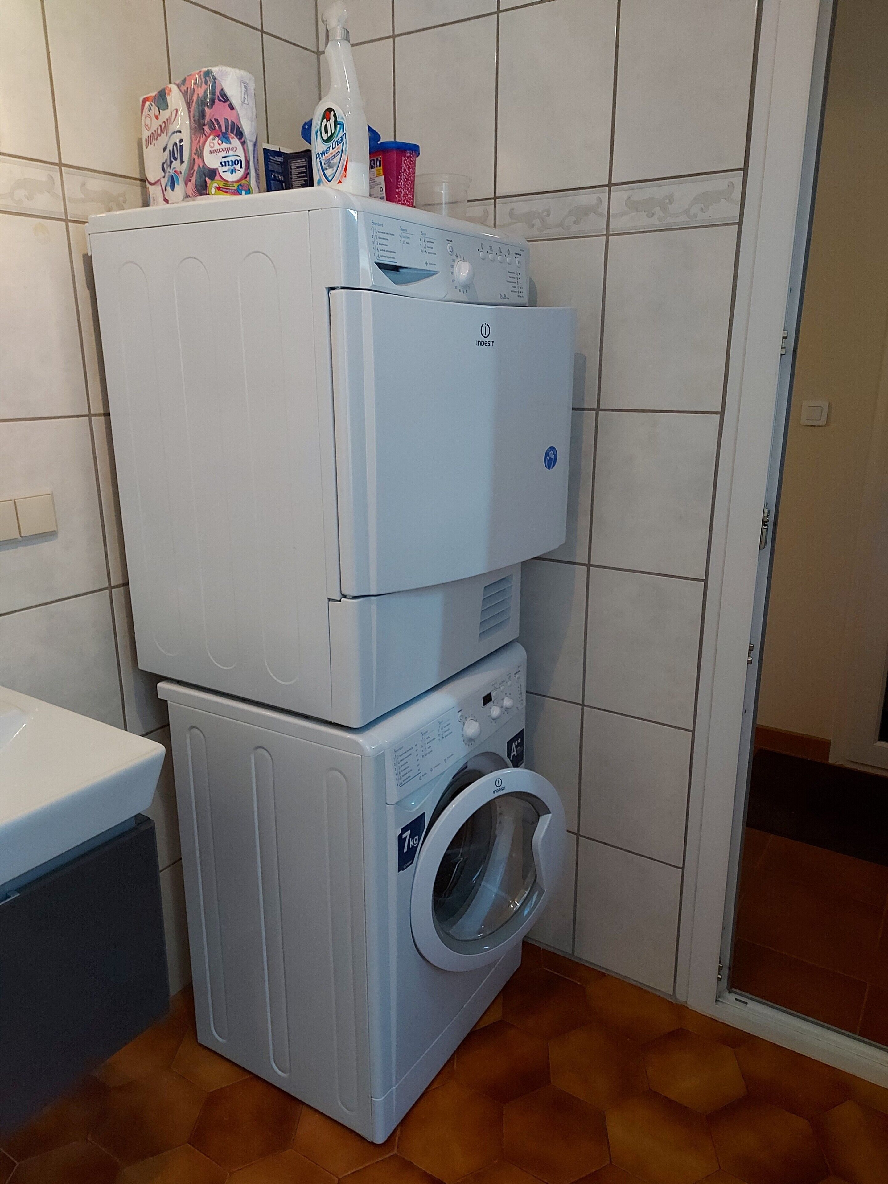 A compact laundry area with stacked appliances, including a washing machine and a dryer, in a room with tiled walls and a tiled floor.