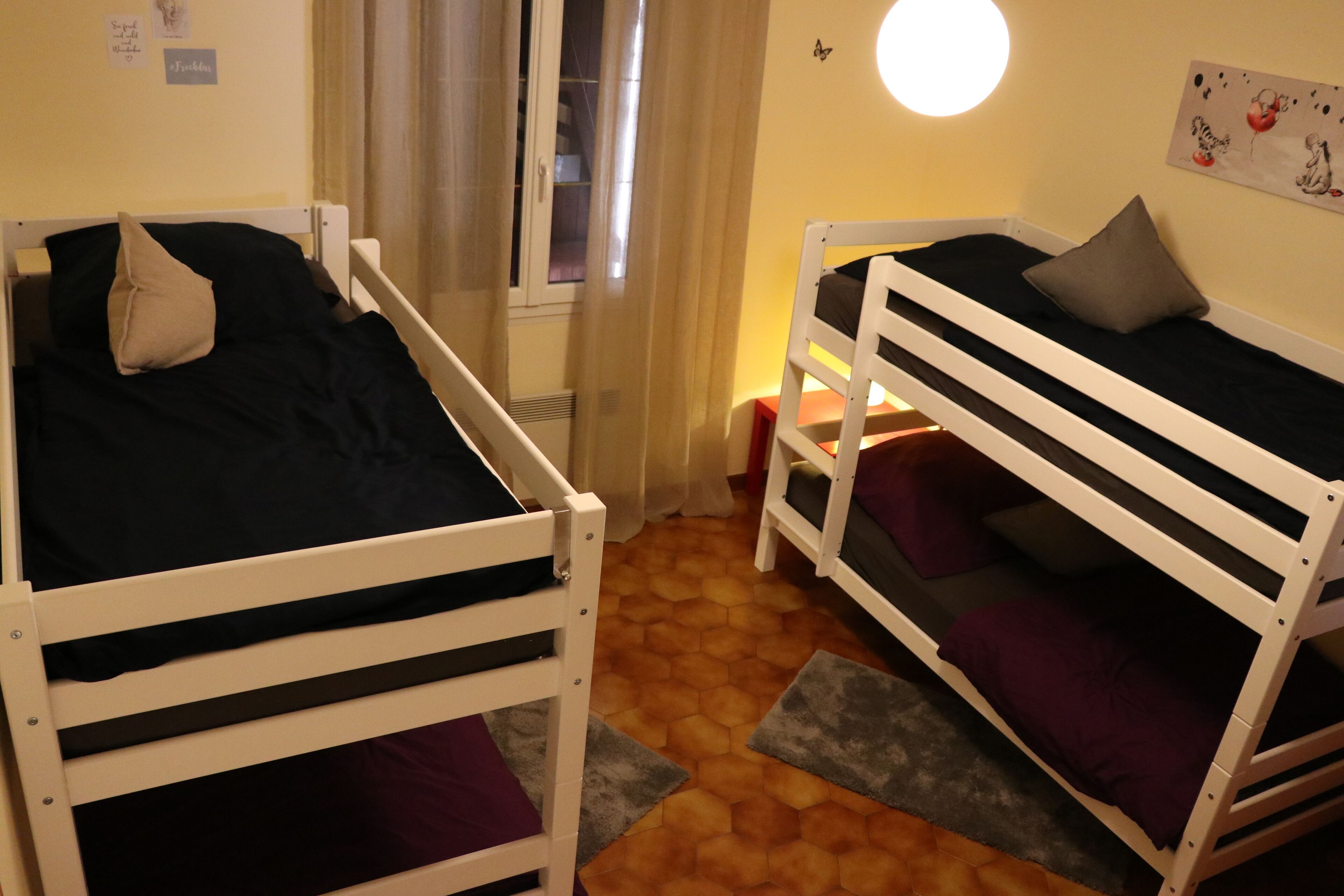Asuntolan huone, jossa on kerrossängyt, ikkuna, jossa on verhot, pyöreä seinävalaisin ja pieni pöytä tuolilla.