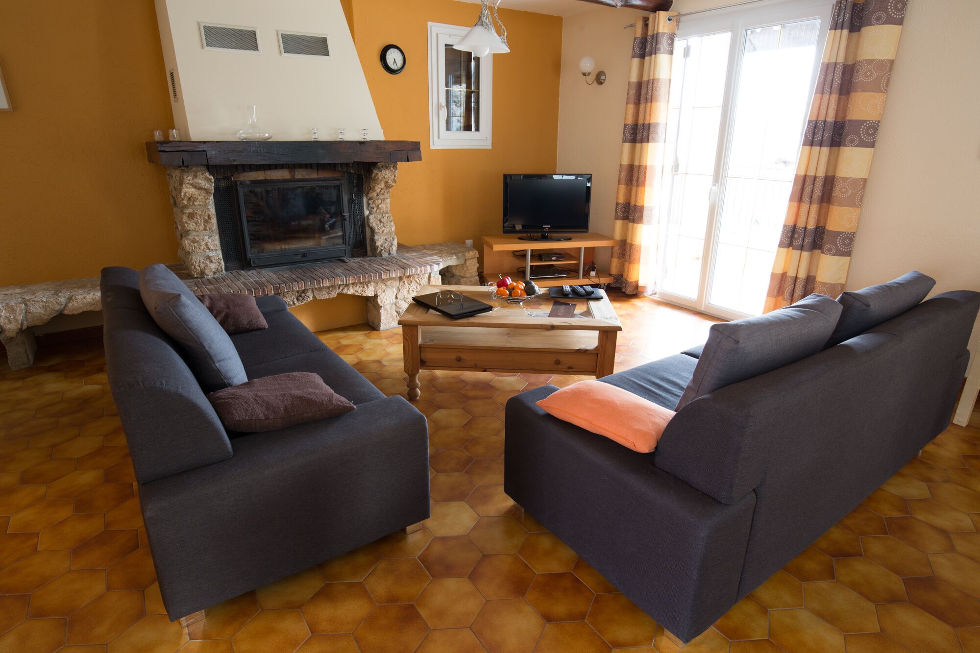 A living room with two grey sofas, a wooden coffee table, a stone fireplace, a television, and a window with curtains.