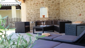 Cómodo espacio al aire libre con mobiliario de mimbre y bancos acolchados, una pared de piedra y un ventanal con vistas a un jardín.