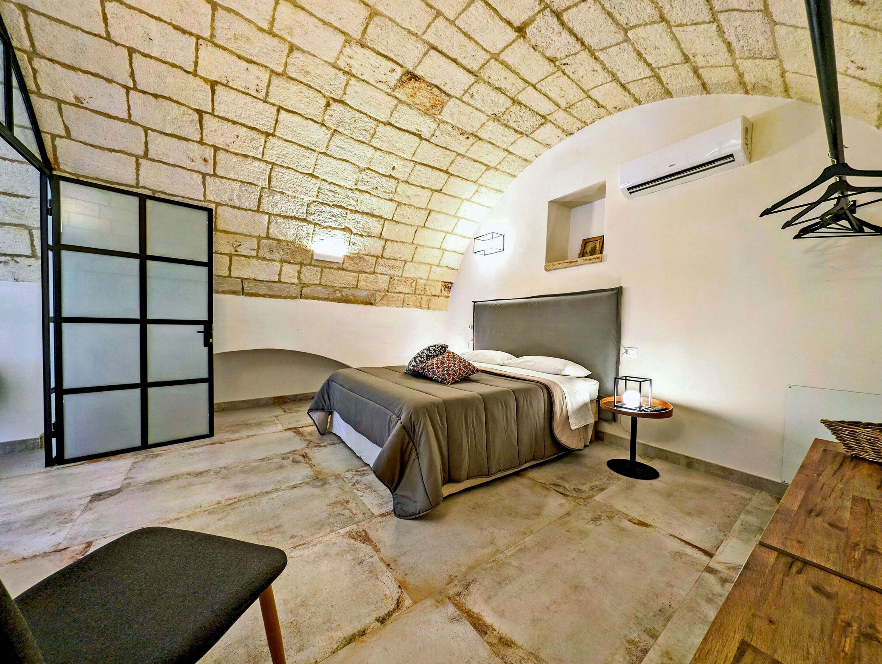 A bedroom with a brick ceiling, a bed with a grey headboard, a bedside table with a lamp, and a chair.