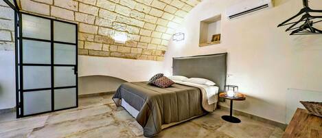 A bedroom with a brick ceiling, a bed with a grey headboard, a bedside table with a lamp, and a chair.