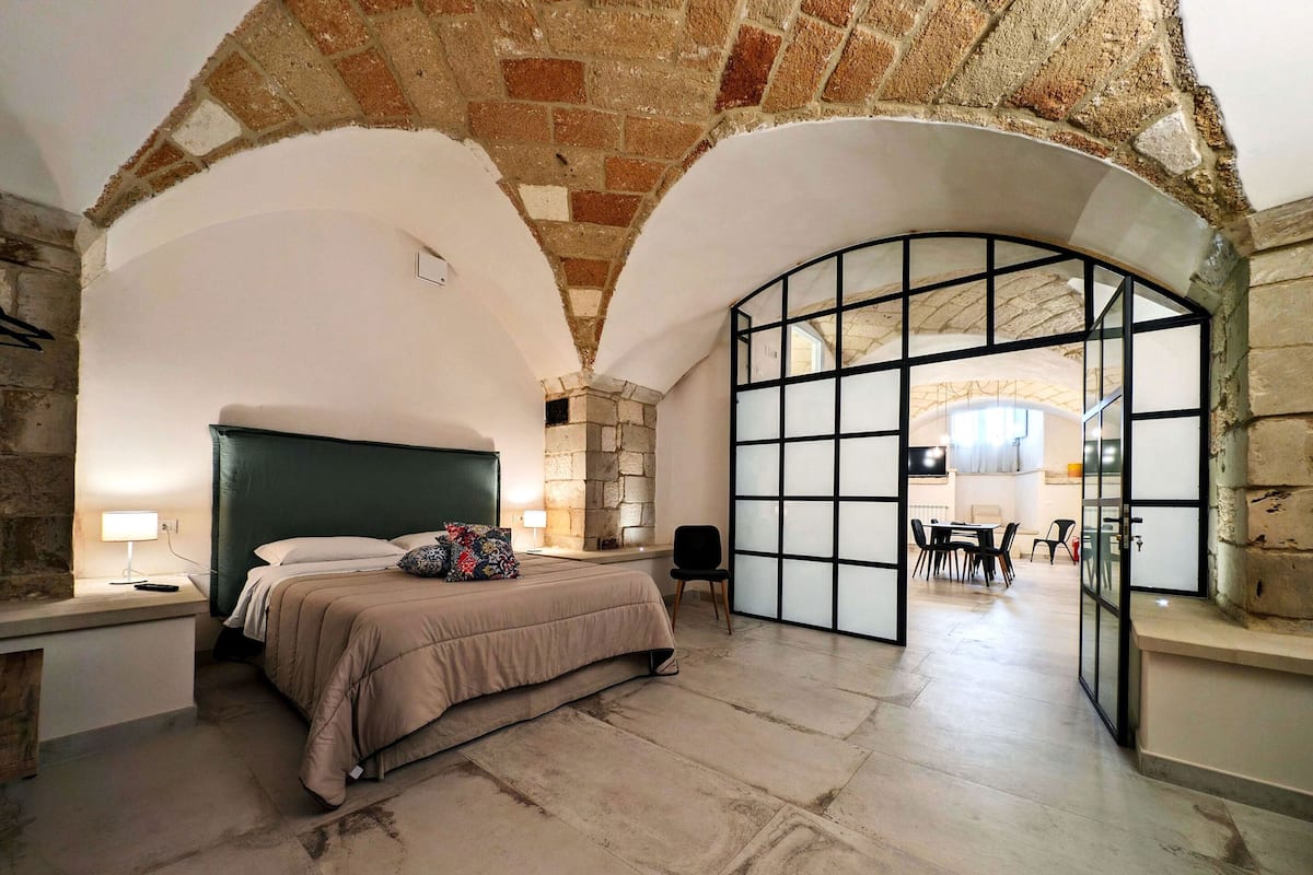 Un dormitorio con chimenea de piedra, una cama grande y vista hacia un comedor a través de un separador de vidrio.