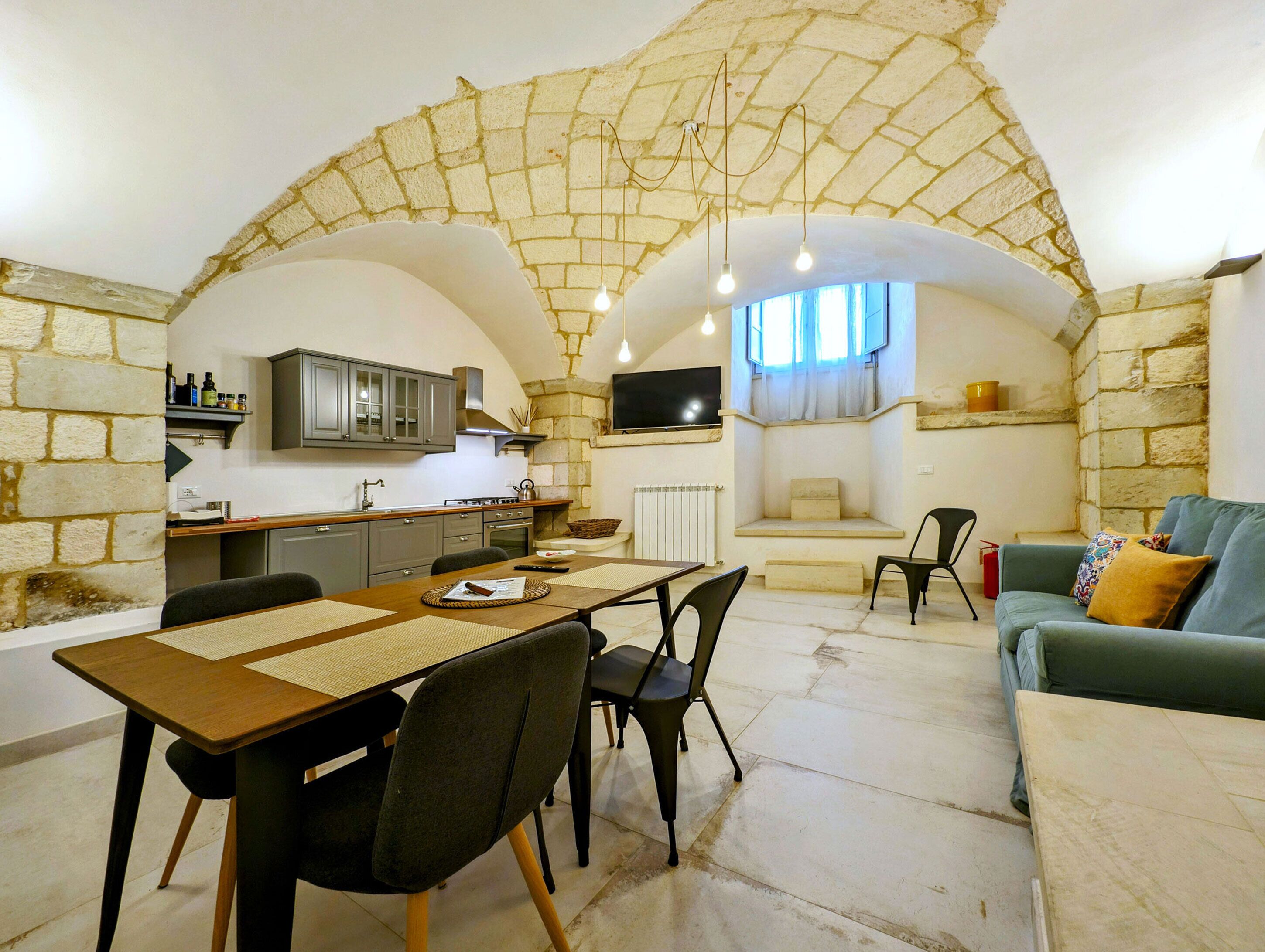 A cozy interior with a stone archway leading to a kitchen and dining area, featuring a stone fireplace, a TV mounted on the wall, and a comfortable seating area with a sofa and chairs.