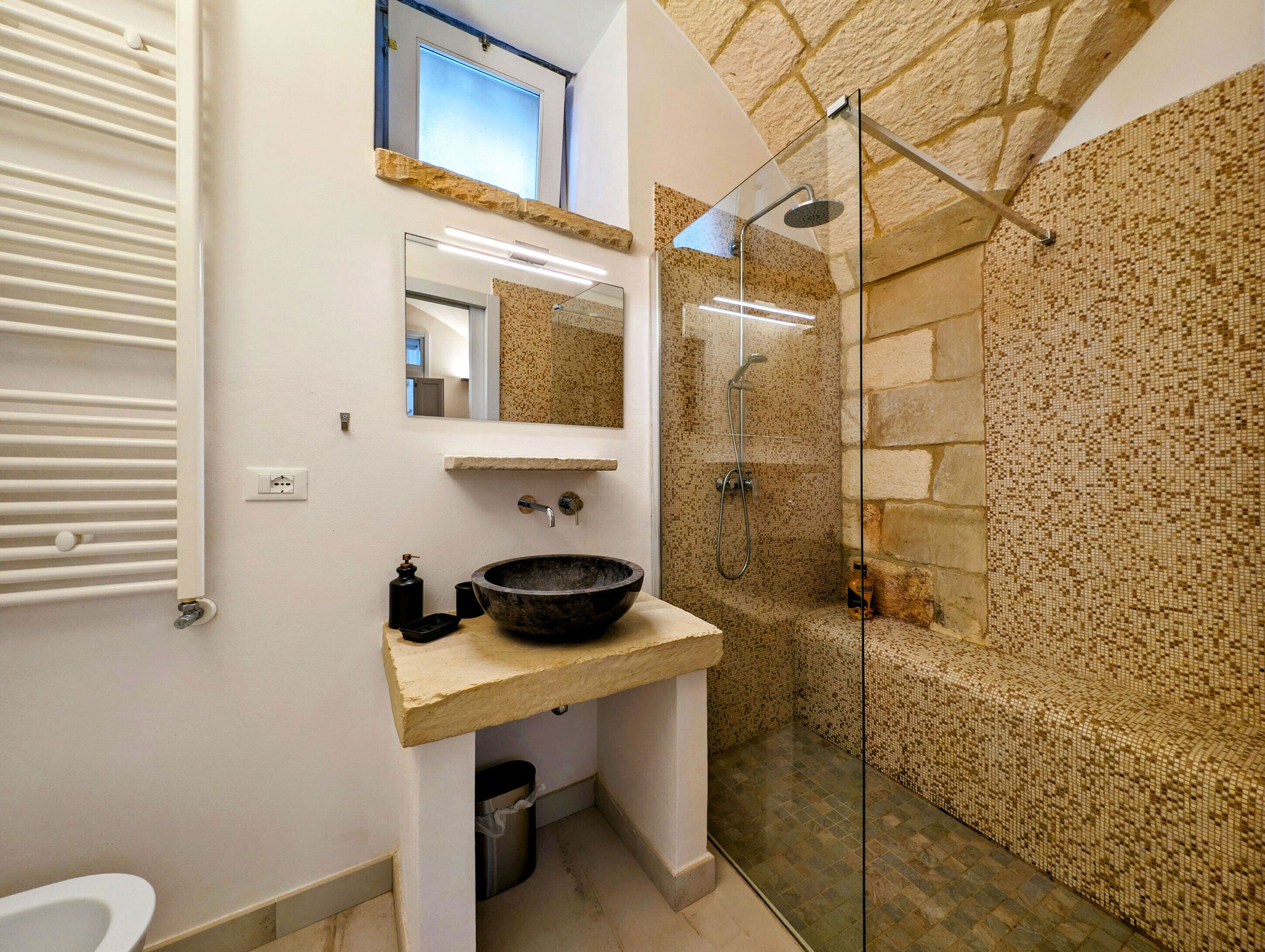 Un baño con pared de piedra, cabina de ducha de vidrio, lavabo espacioso y espejo sobre el lavabo.