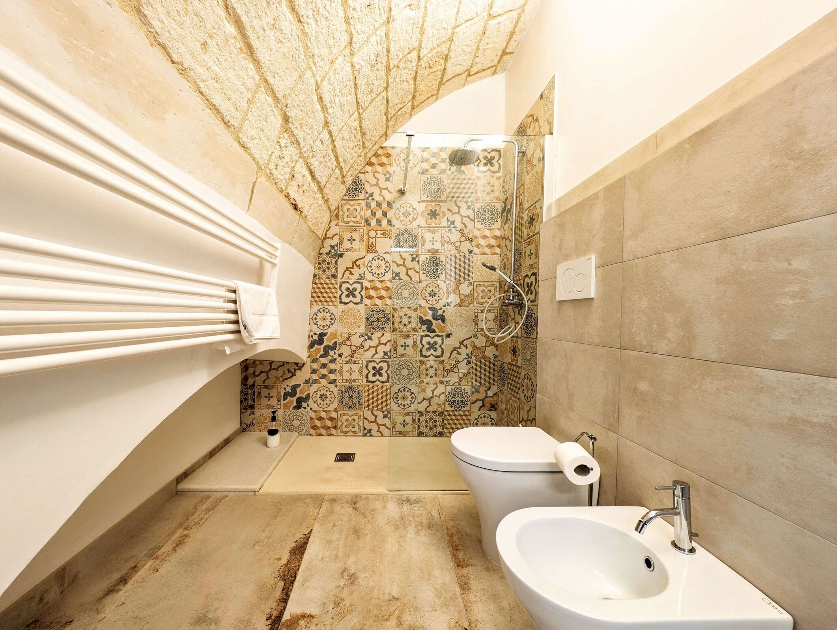 A bathroom with a patterned tile wall, a toilet, a bidet, and a sink.