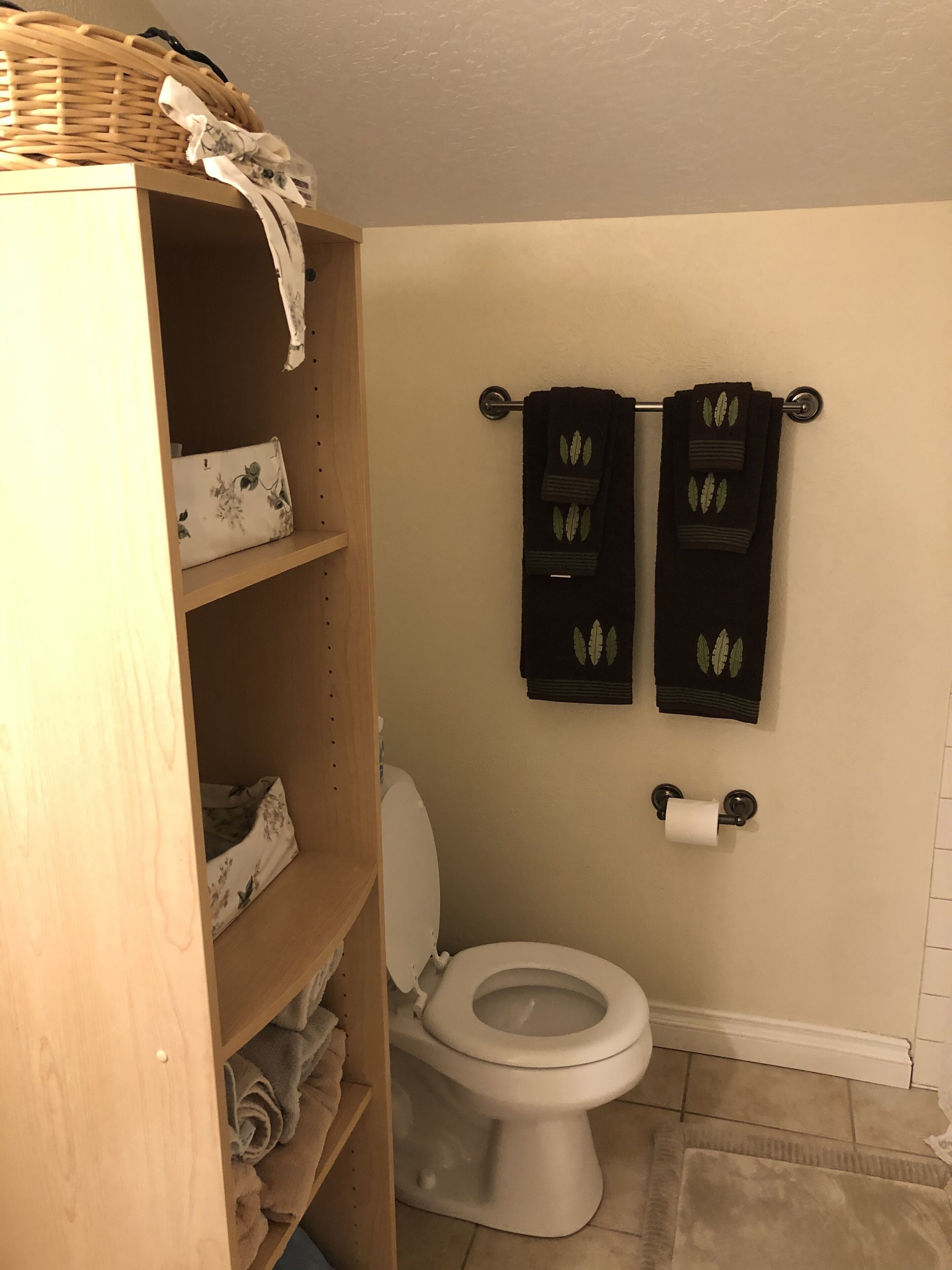 Une salle de bain comprenant des toilettes, un porte-serviettes avec deux serviettes, une étagère en bois avec des serviettes et un panier placé sur le dessus.