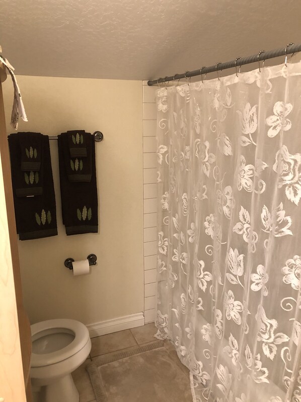 Un baño con un inodoro, dos toallas negras con motivos de hojas, un dispensador de papel higiénico y una cortina de ducha estampada con flores.